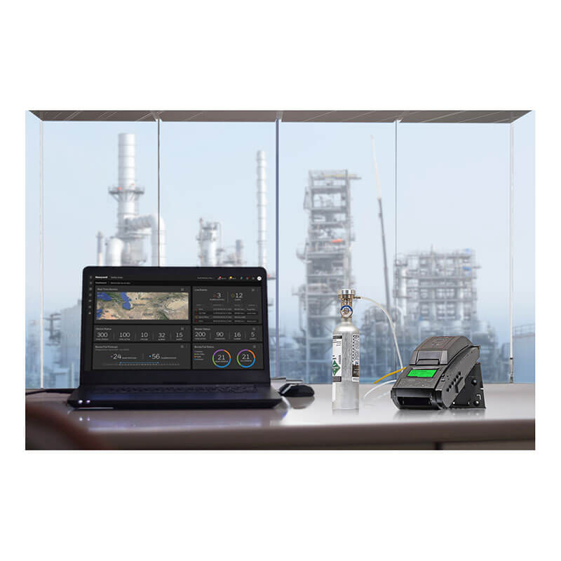 A laptop, a BW Technologies by Honeywell BW Clip4 and IntelliDox Bundle sit on a desk near large windows, overlooking an industrial plant with tall structures in the background.