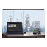 A laptop, a BW Technologies by Honeywell BW Clip4 and IntelliDox Bundle sit on a desk near large windows, overlooking an industrial plant with tall structures in the background.