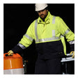 Wearing an NSA DRIFIRE FR Hi-Vis Insulated Jacket JKT-01C3-HV, which is ANSI/ISEA 107 compliant, and a white hard hat, a person moves an orange and white traffic barrel at night.