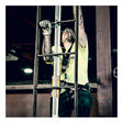 A construction worker wearing a safety harness and gloves climbs an industrial metal ladder equipped with the French Creek Vertical Flexible Cable Climbing System by French Creek Production, looking up while working indoors.