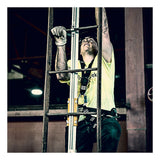 A construction worker wearing a safety harness and gloves climbs an industrial metal ladder equipped with the French Creek Vertical Flexible Cable Climbing System by French Creek Production, looking up while working indoors.