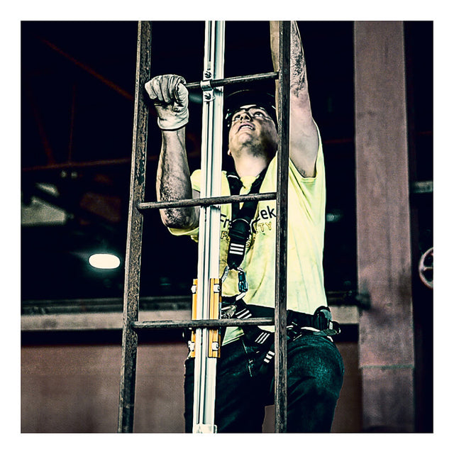 A construction worker wearing a safety harness and gloves climbs an industrial metal ladder equipped with the French Creek Vertical Flexible Cable Climbing System by French Creek Production, looking up while working indoors.