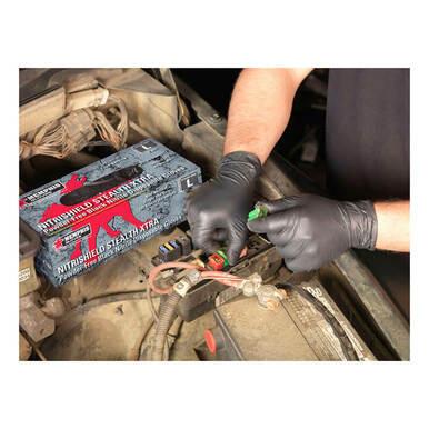 An individual is working under a car hood, adjusting cables on a battery while wearing MCR Safety NitriShield Stealth Extra Gloves 6062, featuring a black textured grip. Nearby, a box labeled "MCR Safety" holds puncture-resistant nitrile gloves. The image highlights hands and automotive components.