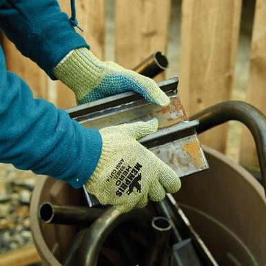 A person wearing MCR Safety's Hero Kevlar Stainless Steel Cut Protection Gloves 93867, offering ANSI Cut Level A4 protection, holds scrap metal pieces over a large brown container, with a wooden fence in the background.