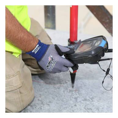 Kneeling on a concrete surface, a person wearing MCR Safety's MCR Ninja BNF Evolution with Ingenia 15g N96051 gloves operates a surveying device enhanced with NFT coating. The device is mounted on a red pole, and the individual is dressed in a high-visibility shirt and beige pants.