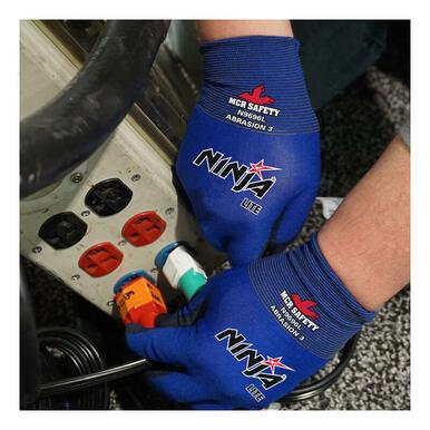 A person wearing blue MCR Safety Ninja Lite Work Gloves N9696, featuring a PU Coated Palm, is seen plugging a power cord into a socket. The gloves prominently display the "MCR Safety" and "Ninja Lite" logos. In the background, part of an electrical panel with red and white outlets can be seen, emphasizing the gloves' practicality for precision tasks.