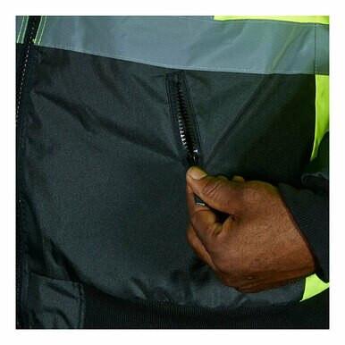 A hand zips up the pocket of a dark, two-tone bomber jacket. The MCR Safety Luminator Two Tone Value Bomber Jacket VBBCL3L is designed with high visibility in mind, featuring bright yellow and gray accents that are perfect for standing out in any setting.