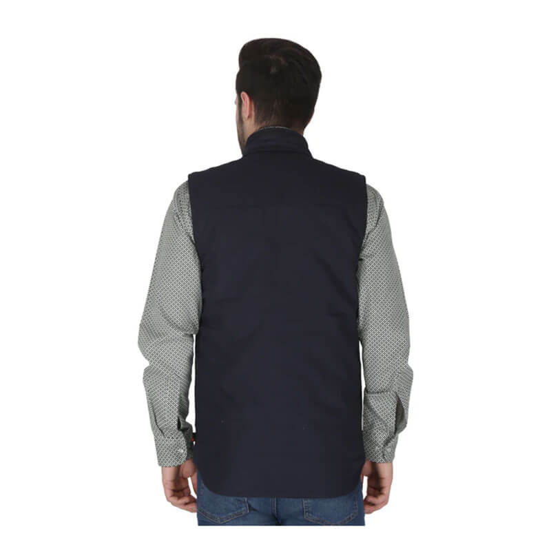 A man stands with his back to the camera, wearing a ForgeFR Forge FR Canvas Duck Vest MFRVST-001 over a light grey patterned long-sleeve shirt and blue jeans. The plain white background highlights this flame-resistant workwear.