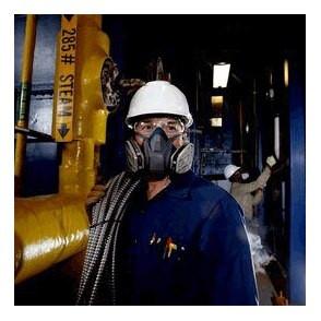 A worker in a white helmet and a 3M 6500QL Quick-Release Half Face Respirator Mask stands in an industrial setting, holding cables. In the background, another worker tends to equipment. Nearby, a yellow steam pipe with a quick-release feature and control valve is visible.