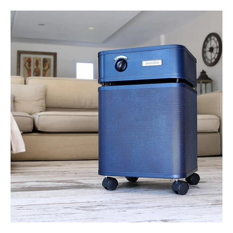 An Austin Air Systems HealthMate Plus Machine B450 air purifier in blue, equipped with wheels, is positioned on a wooden living room floor. It features advanced medical-grade HEPA filtration for effective formaldehyde removal, with a beige sofa and wall clock visible in the background.
