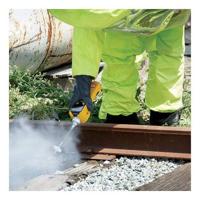 A person wearing a bright yellow protective suit and dark gloves is using a power tool, possibly with guidance from the RAE Systems MiniRAE 3000 PID, to apply a substance or perform maintenance on a rusty metal rail. Smoke or mist is visible around the work area, with small plants in the background.