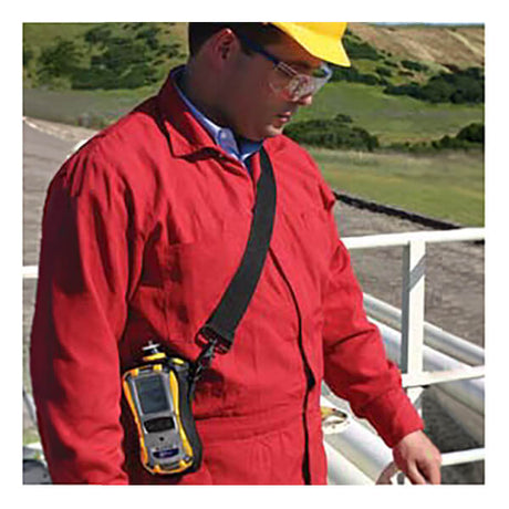 A person in a red coverall, yellow hard hat, and safety glasses stands outdoors with a RAE Systems MultiRAE Lite Monitor (Pump and Non-Wireless PGM-6208) clipped to their suit for gas detection; green hills are visible behind them.