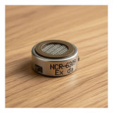 A close-up shows an RKI Instruments Replacement LEL Sensor NCR-6309, with a mesh-covered top and metallic body, resting on a wooden surface. It's commonly used as an RKI LEL replacement in gas monitors such as the GX-3R Pro.