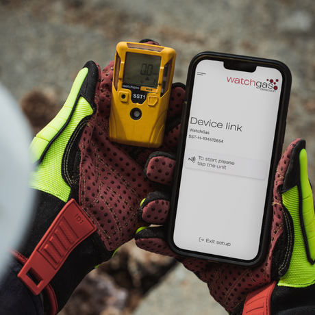 Person holding a yellow gas detector and a smartphone with 'watchgas' app.