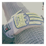 A gloved hand rests on a roll of metal wire mesh. The glove, part of the MCR Predator Hi-Vis Mechanics Gloves with HyperMax (PD6951) from MCR Safety, features gray material with yellow and blue padding and is specifically designed for cut resistance—making it ideal for handling sharp materials. Sunlight washes over the scene, producing a bright, slightly overexposed effect.