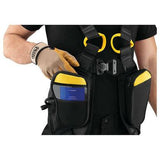 A person wearing the Petzl Newton Easyfit International Harness C073FA in black, featuring automatic buckles and padded shoulder straps, is adjusting its strap. The harness includes two distinctive black and yellow pouches, with one containing a blue item. A tan glove covers one hand.