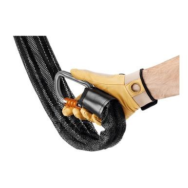 A hand wearing a tan work glove holds the Petzl JAG System P044AA0, a heavy-duty black metal carabiner with sealed ball bearings, which is attached to a braided black rope.