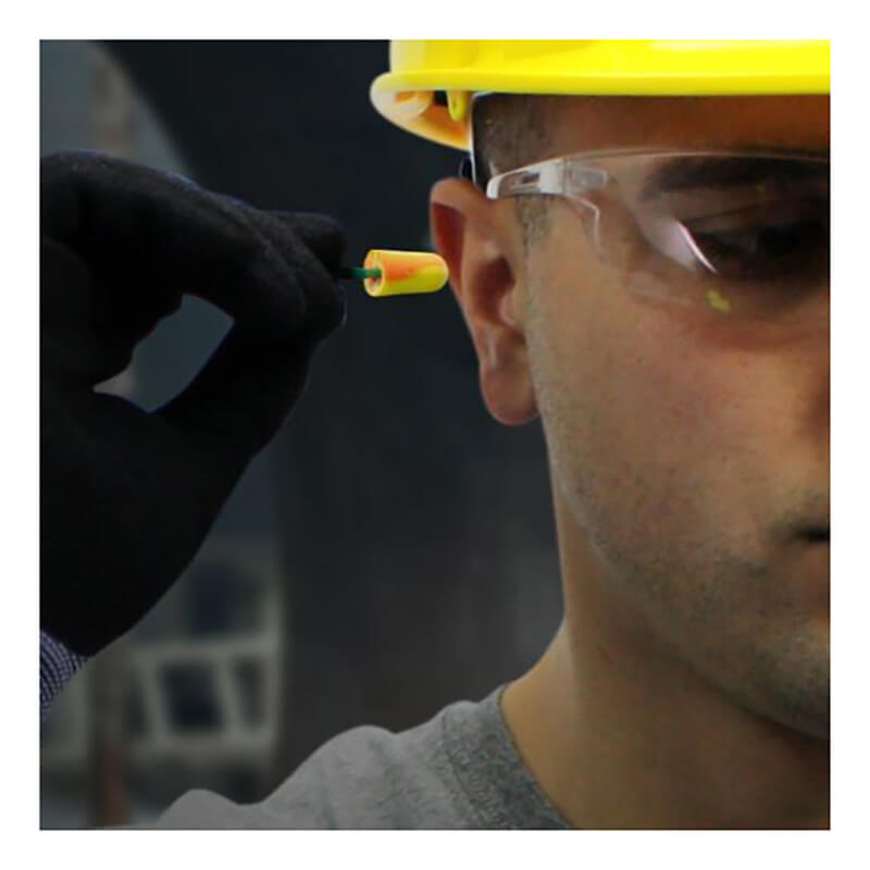 A person wearing a yellow hard hat and safety glasses uses a gloved hand to insert a PIP Pinchfit BioSoft Disposable Foam Ear Plug PF-1, highlighting the importance of hearing protection on the job.