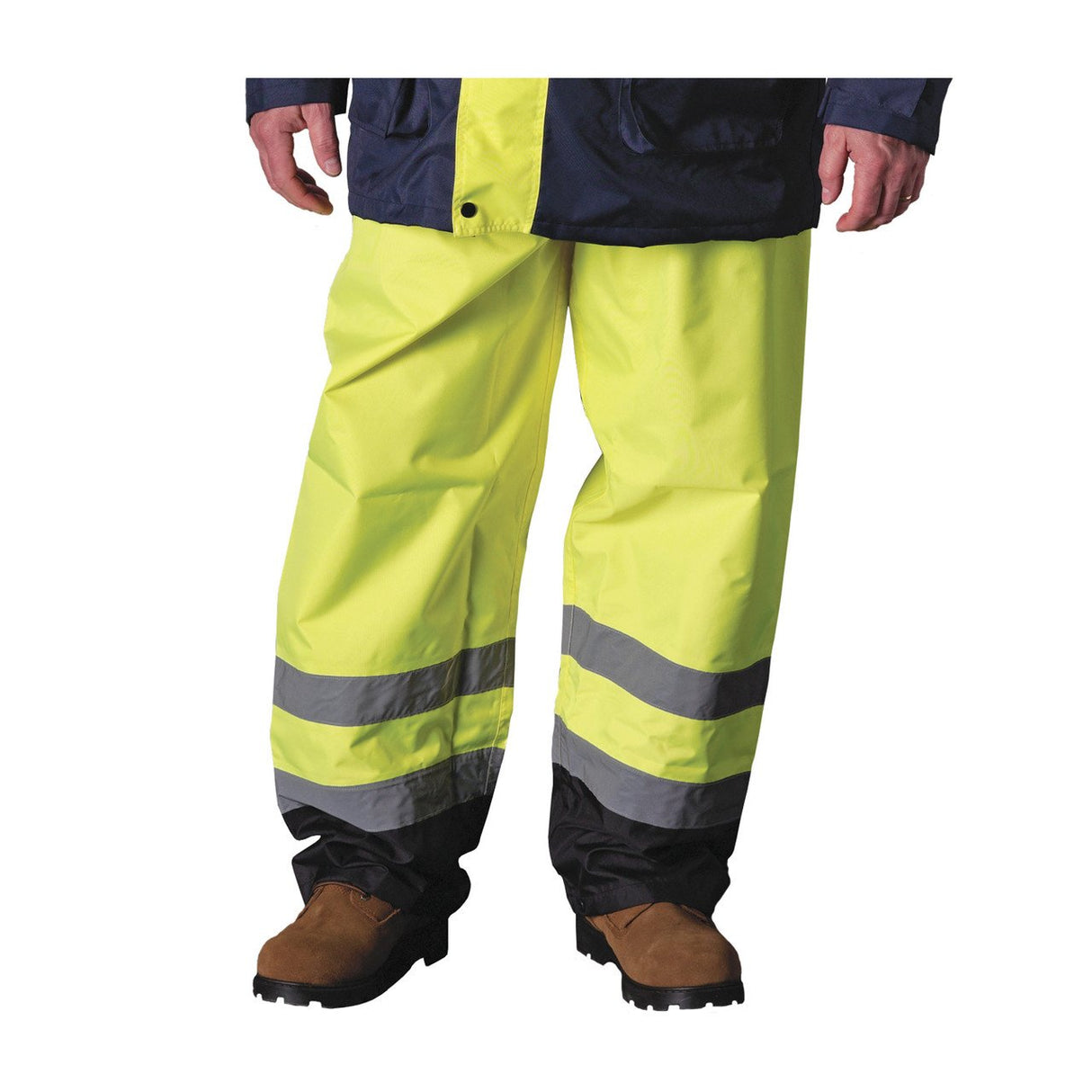 Dressed in PIP Class E Value Black Trim Over Pant 318-1757, made from waterproof polyester and featuring reflective gray and black stripes near the cuffs, the person complements their attire with brown work boots. Their partially visible dark jacket completes a perfect hi-visibility ensemble for safety.