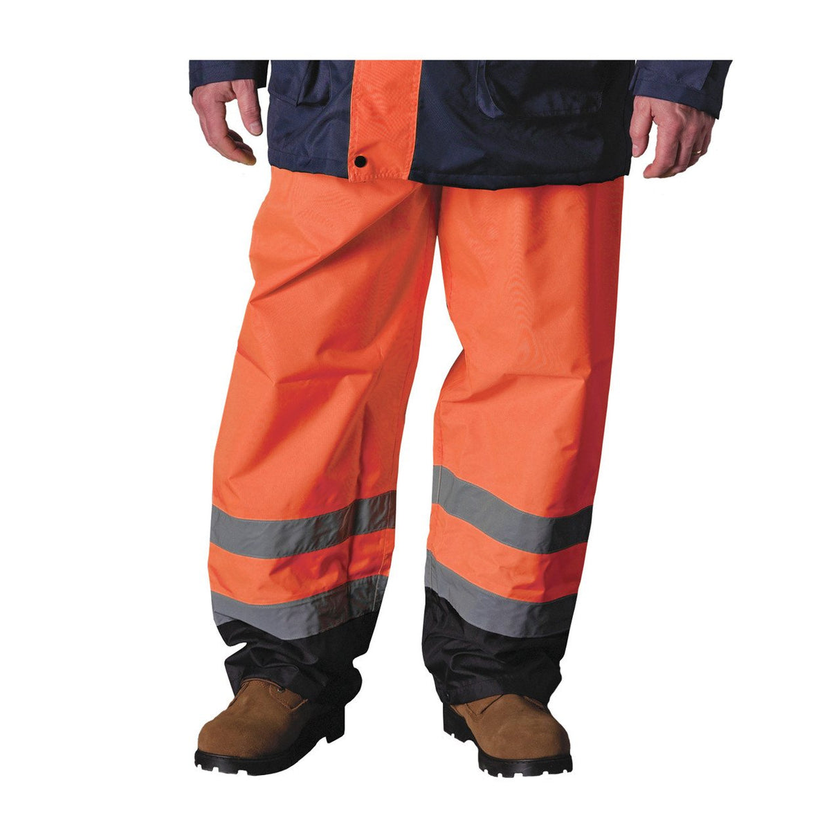 A person wearing PIP Class E Value Black Trim Over Pants 318-1757, which are made from waterproof polyester in orange with gray stripes for enhanced visibility, stands in brown work boots. Their upper body is covered by a dark jacket, while the image emphasizes the hi-visibility PIP over pants covering their lower half.