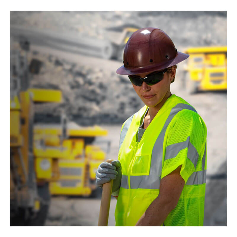 A construction worker wearing a PIP Wolfjaw Full Brim Smooth Dome Hard Hat w/Fiberglass 280-HP1481R, sunglasses, neon yellow safety vest, and gloves holds a tool, with industrial equipment and a blurred construction site in the background.
