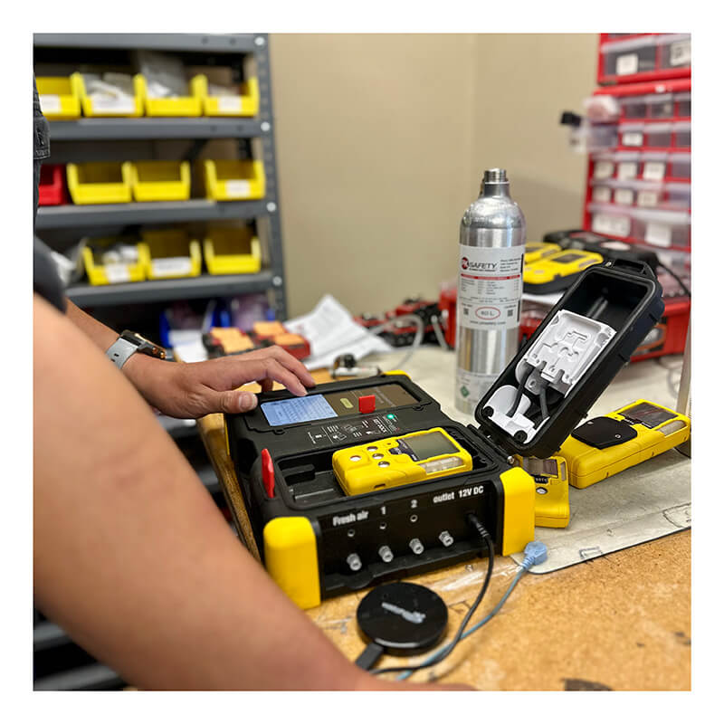 A person's hand is shown calibrating a WatchGas monitor on docking station at a work desk with gas detection components surrounding them including a silver gas cylinder in the background.