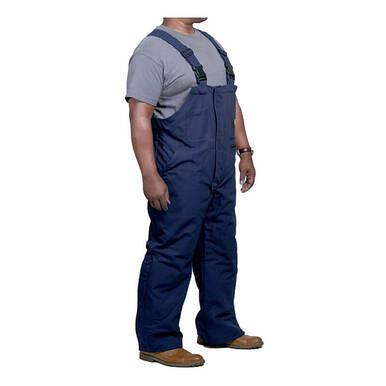 A person wearing GRIT FR Insulated Bib Overalls in dark blue with quilt-lined modacrylic insulation, paired with a gray t-shirt and brown work boots, stands against a white background.