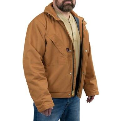 A person is wearing a GRIT FR Insulated Field Jacket in tan, crafted by GRIT FR Workwear from fire-resistant cotton duck canvas over a beige shirt and blue jeans. This quilt-lined insulated jacket features zip and button closures along with practical front pockets, emphasizing its durable design.