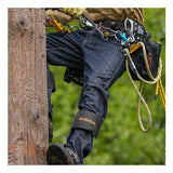 Wearing NSA’s DRIFIRE Slim Straight Leg FR Jean PNT-JSS01 along with other safety gear and climbing equipment, a person scales a wooden utility pole surrounded by greenery in the background.