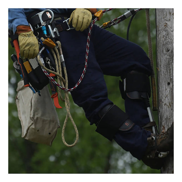 Wearing the NSA DRIFIRE Range Flex Pro Relaxed FR Work Pant in navy and arc-rated safety gear, a utility worker ascends a wooden pole with climbing equipment, tools hanging from their belt and ropes secured, set against a blurred green background.
