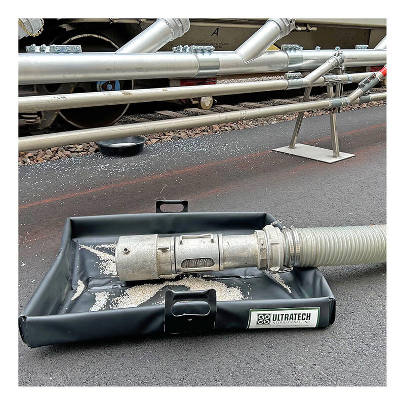 A metal hose connector sits in a black UltraTech Pellet Pond 8859 by UltraTech on asphalt, serving as part of a pellet containment system to capture scattered plastic granules during industrial piping work.