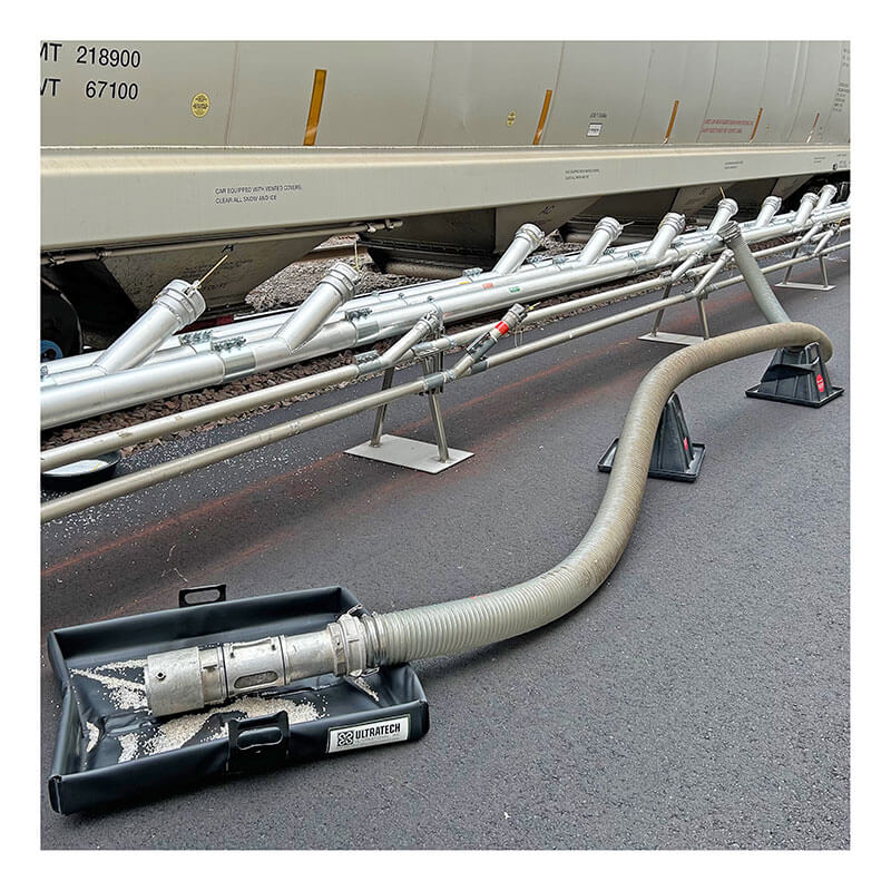 The UltraTech Pellet Pond 8859 by UltraTech is placed under connections where a large industrial hose meets metal piping beside a train car, providing spill and plastic pellet containment on an asphalt surface.