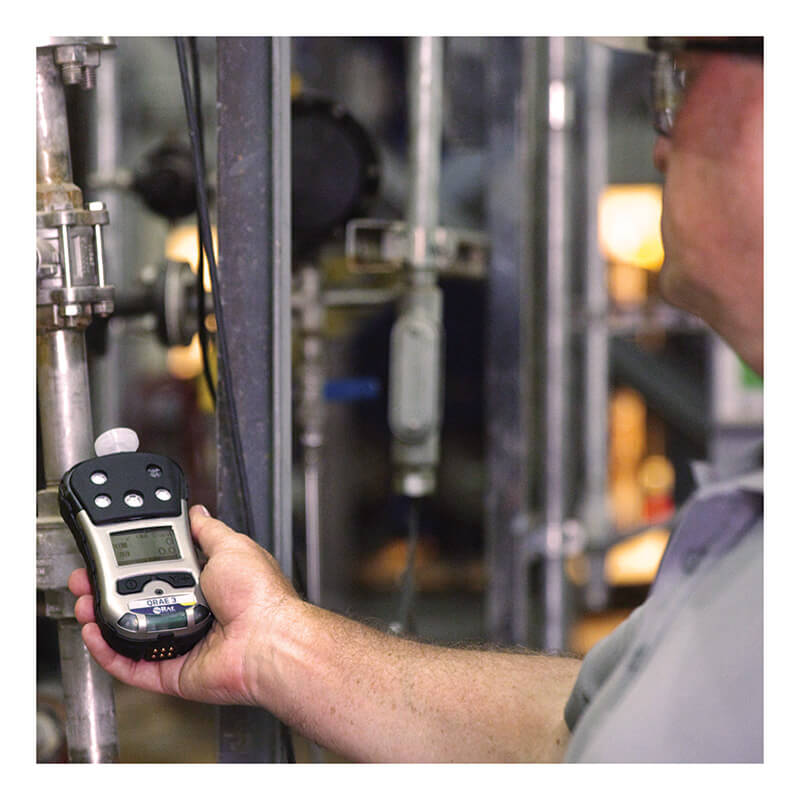 A person uses the RAE Systems PGM-2500 QRAE 3 Wireless Pumped Multi-Gas Monitor M020-11211-111 near pipes and industrial equipment to check gas levels in an industrial setting, with their face out of focus.