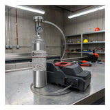 An RKI Instruments SDM-3R Calibration Station Bundle is connected via tubing to a metal gas cylinder with a pressure gauge, all set up on a stainless steel workbench in an industrial lab. The area is equipped with calibration tools and surrounded by shelves, various equipment, and safety helmets in the background.