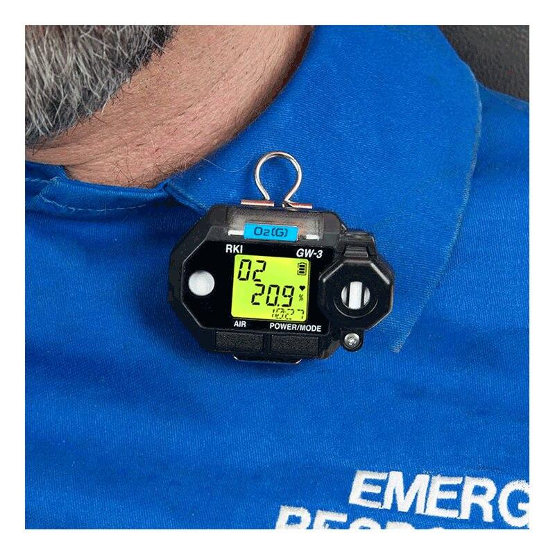 A close-up of a person wearing a blue shirt with "EMERG" partially visible, featuring an RKI Instruments GasWatch 3 O2 Single Gas Monitor 72-0012 clipped to the fabric. The device's yellow screen displays various numbers, indicating continuous operation and vital readings.