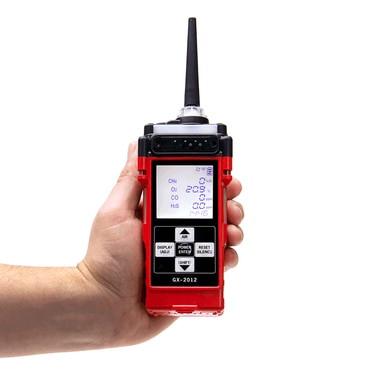 A hand holds the RKI Instruments GX-2012 5-Gas Detector, a red handheld device equipped with advanced micro-sensor technology and a digital display showcasing readings for oxygen, carbon monoxide, hydrogen sulfide, and combustible gases. It is perfect for confined space entry and includes several buttons along with a black antenna.