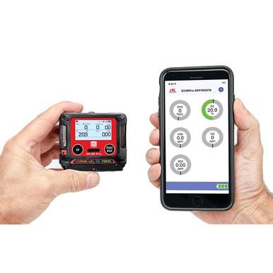 In a confined space, two hands are holding devices: one is the compact RKI GX-3R Pro 5 Gas Confined Space Monitor from RKI Instruments, featuring a red display with "209" and "20.9," while the other is a smartphone using wireless communication to show an app with circular graphics displaying similar numbers.