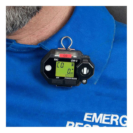 In a close-up shot, a person is seen wearing a blue uniform with the RKI Instruments GasWatch 3 CO Gas Personal Monitor 73-0023 clipped on. The device includes an IP-66/68 rated gas sensor that clearly displays "CO" with a carbon monoxide level reading of 0 ppm on its digital screen.