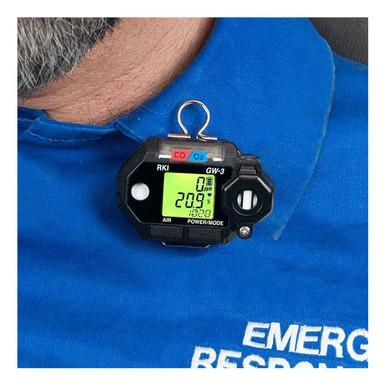 The RKI Instruments GasWatch 3 CO/O2 Gas Personal Monitor 73-0026, clipped to blue emergency responder clothing, displays gas readings on its screen. The numerical values indicate levels for oxygen and carbon monoxide, utilizing its CO/O2 sensor.