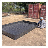 A worker in gray attire and a helmet sets up an UltraTech Concrete Washout Berms - RPE Model by UltraTech, a large black reinforced polyethylene berm, on dirt near shipping containers and trees.