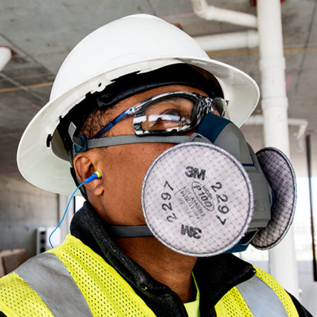 Person wearing a hard hat, safety glasses, and a 3M respirator mask in an industrial setting.