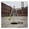 The French Creek Production 7' Tripod Rescue System S50G-7 stands over an open manhole in an empty industrial courtyard, yellow and silver against a backdrop of brick buildings and smokestacks.