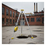 The French Creek Production 7' Tripod Rescue System S50G-7 stands over an open manhole in an empty industrial courtyard, yellow and silver against a backdrop of brick buildings and smokestacks.