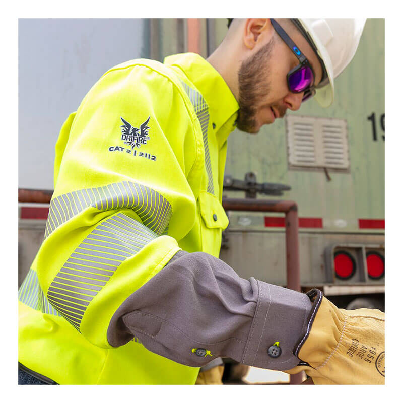 A worker in a white hard hat, sunglasses, gloves, and the NSA DRIFIRE FR Hi-Vis Lightweight Work Shirt Class 3 SHRTS1C3 operates equipment near a truck. The flame resistant shirt features a logo and text on the sleeve.