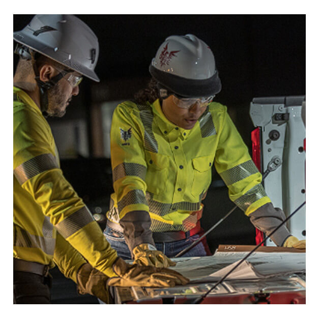 Two construction workers in safety gear and helmets review blueprints on a table, both wearing NSA DRIFIRE FR Premium Hi-Vis Vented Shirt SHRTVTGVC3 as they focus intently on the plans under work lights, likely during a nighttime project.
