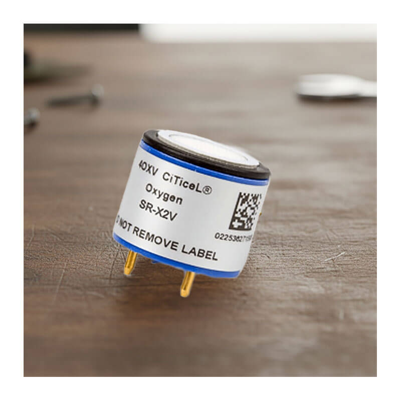 A close-up of a Replacement O2 Sensor for GasAlert Monitors SR-X2V by BW Technologies by Honeywell, showing its cylindrical shape, metal pins, and a warning label stating “DO NOT REMOVE LABEL,” resting on a wooden surface.