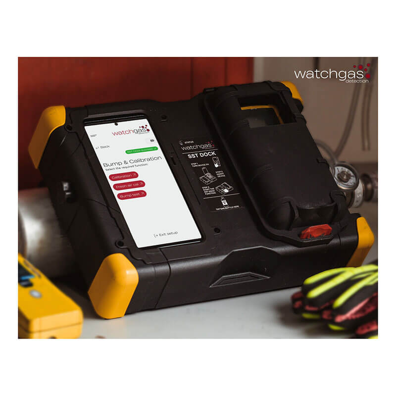 A black and yellow WatchGas gas detector with a digital screen sits on a workbench next to safety gloves, tools, and a WatchGas Docking Station w/Touchscreen SST-DOCK for efficient calibration.