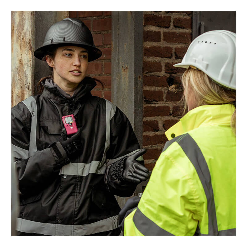 Two workers in safety gear and hard hats talk. One, wearing a black jacket, gestures while holding a pink WatchGas SST1 3 Year H2S Gas Detector (model SST1-H-36-P) by WatchGas; the other, in a yellow jacket, listens with back to camera. A brick wall and concrete structure are visible behind them.