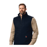 A bearded man wears the ForgeFR Forge FR Canvas Duck Vest MFRVST-001, a black flame-resistant vest, over a beige long-sleeve shirt while standing against a plain white background.