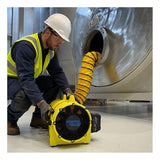 A worker in safety gear sets up a Euramco RAMFAN UB20 18VDC and Quick-Couple Canister ventilator near a large metal tank, connecting its flexible yellow duct to an opening in the tank to ensure effective jobsite and confined-space ventilation.